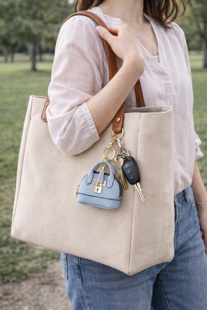 Person holding a beige tote bag with a small blue petite leather purse bag charm and keys attached, outdoors.