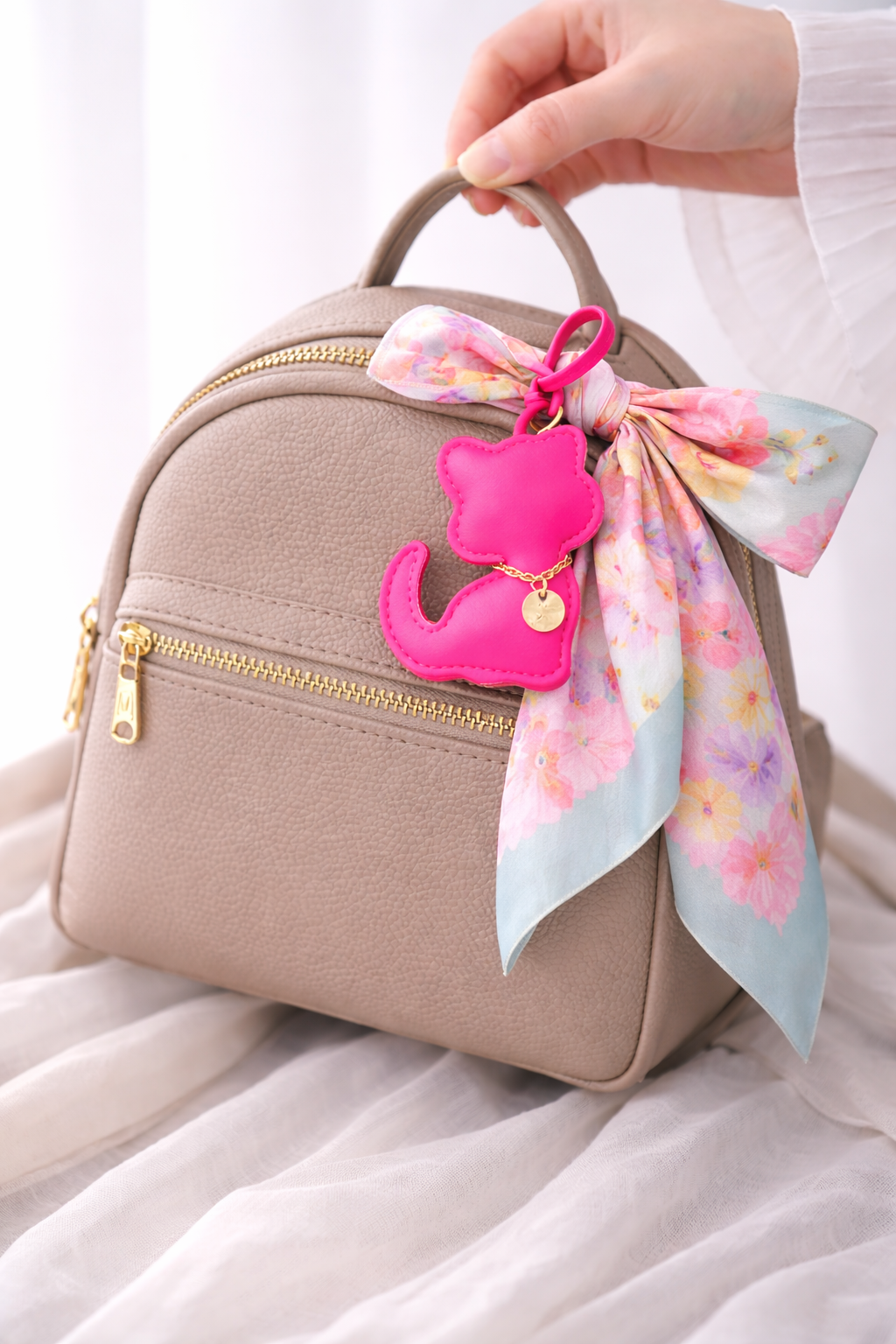Beige handbag with a pink kitten bag charm and colorful scarf on a white background.