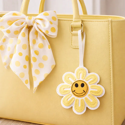 A Yellow Smiles in Bloom Daisy Bag Charm on a yellow purse with scarf on table. 