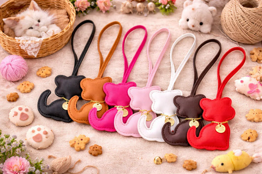 7 different colored Cozy Kitty Handmade Leather Kitten Bag Charms with leather straps in a row on a blanket with other Catt accessories.  
