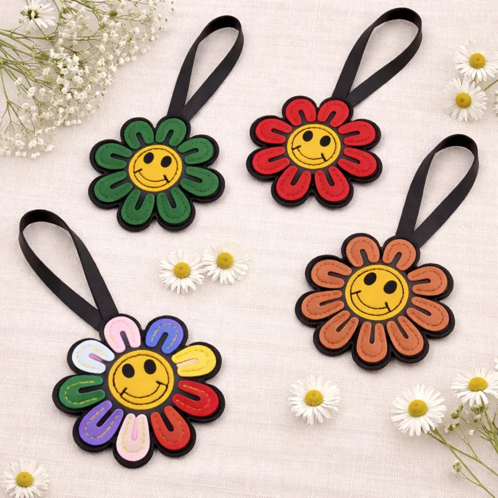 5 different colored Smiles in Bloom Daisy Bag Charms in a circle on table with flowers. 