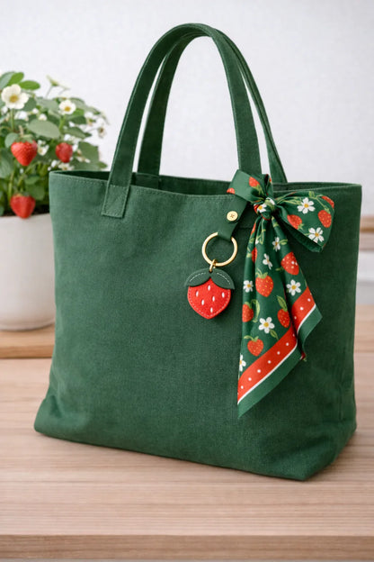 Little Orchard Strawberry Fruit Bag Charm with leather strap on green bag with scarf sitting on table. 