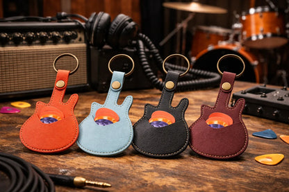 4  leather guitar pick holder key chains showing standard guitar picks tucked inside the secure sleeve, with music related accessories on table. 