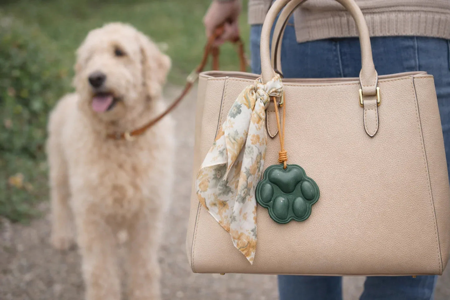 A Woman walking a dog in a park with an Everyday Companion Green Dog Paw Bag Charm with leather strap on pink purse and scarf. 