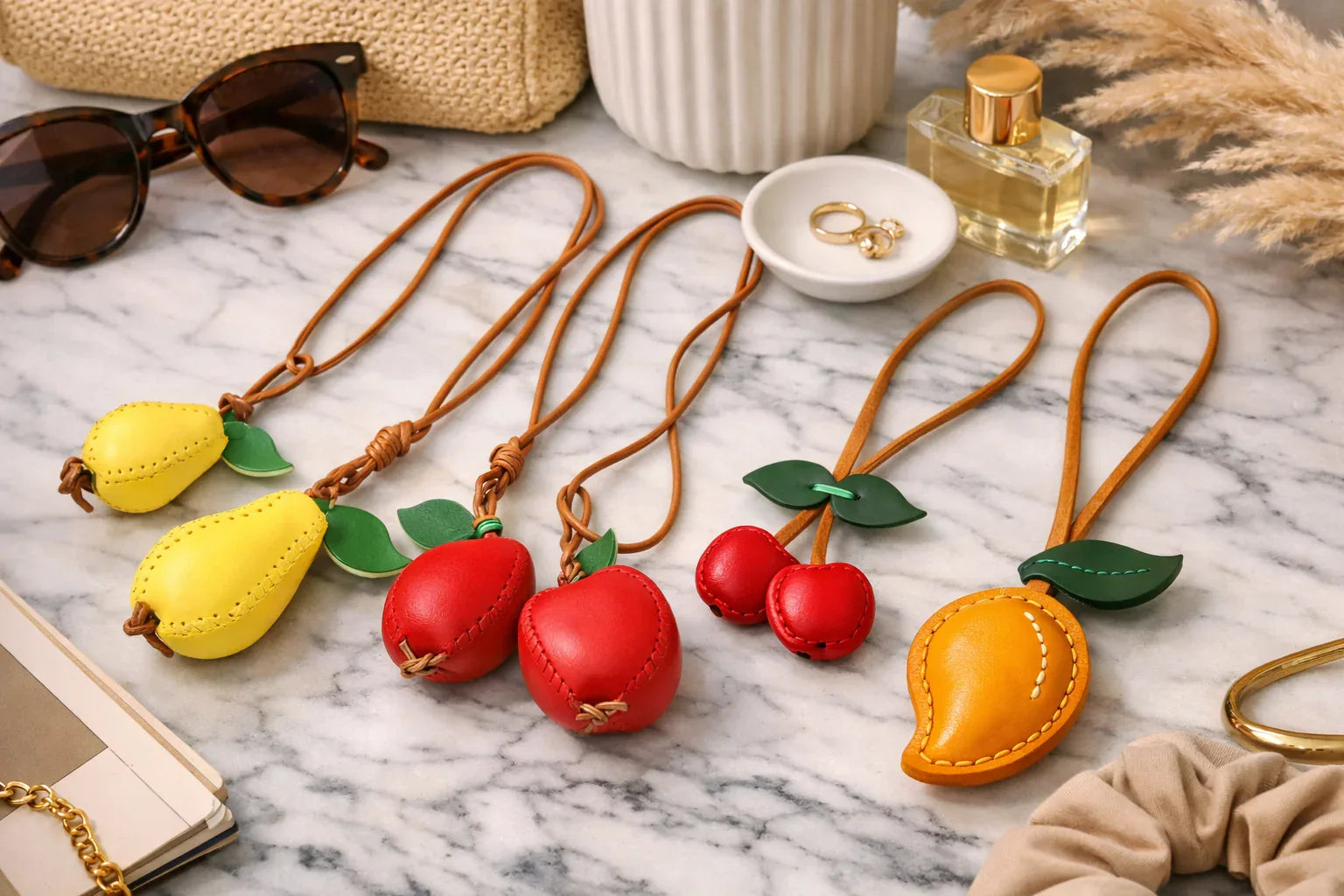 4 different Orchard Luxe Leather Fruit Bag Charms (Mango, Cherry, Pear, Apple)  with leather strap on marble table. 