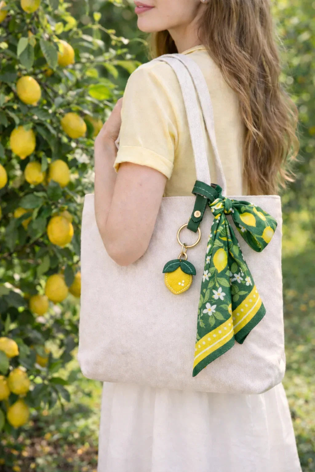 Woman outside in lemon orchard, carrying white tote bag with scarf with Little Orchard Lemon Fruit Bag Charm with leather strap on it. 