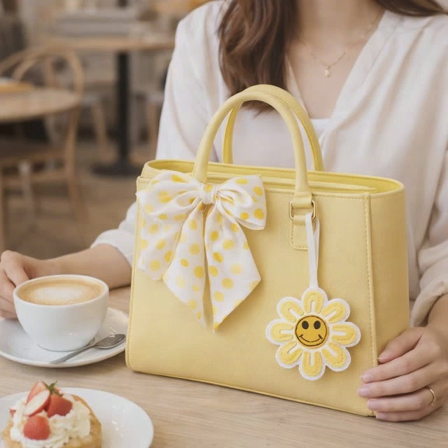 Video of Woman holding a Yellow Smiles in Bloom Daisy Bag Charm on a yellow purse with scarf having coffee. 