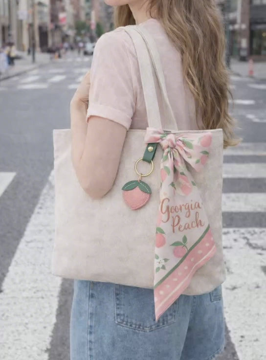 Video of woman walking in city with tote bag with Little Orchard Peach Fruit Bag Charm with leather strap connected. 